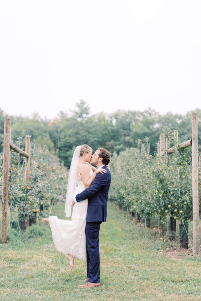 Bride and groom in the middle of an orchard share a kiss as the groom liftss bride up