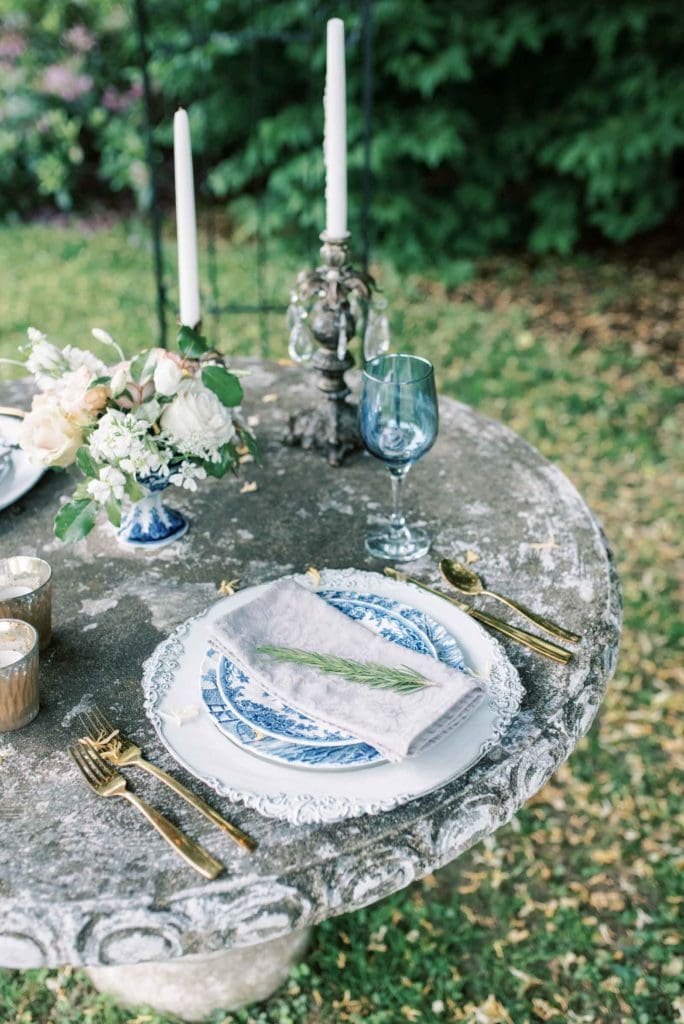 Close-up shot of a round table the plate with a napkin and plant placed on top of it, along with a blue wine glass, utensils, candles and flowers