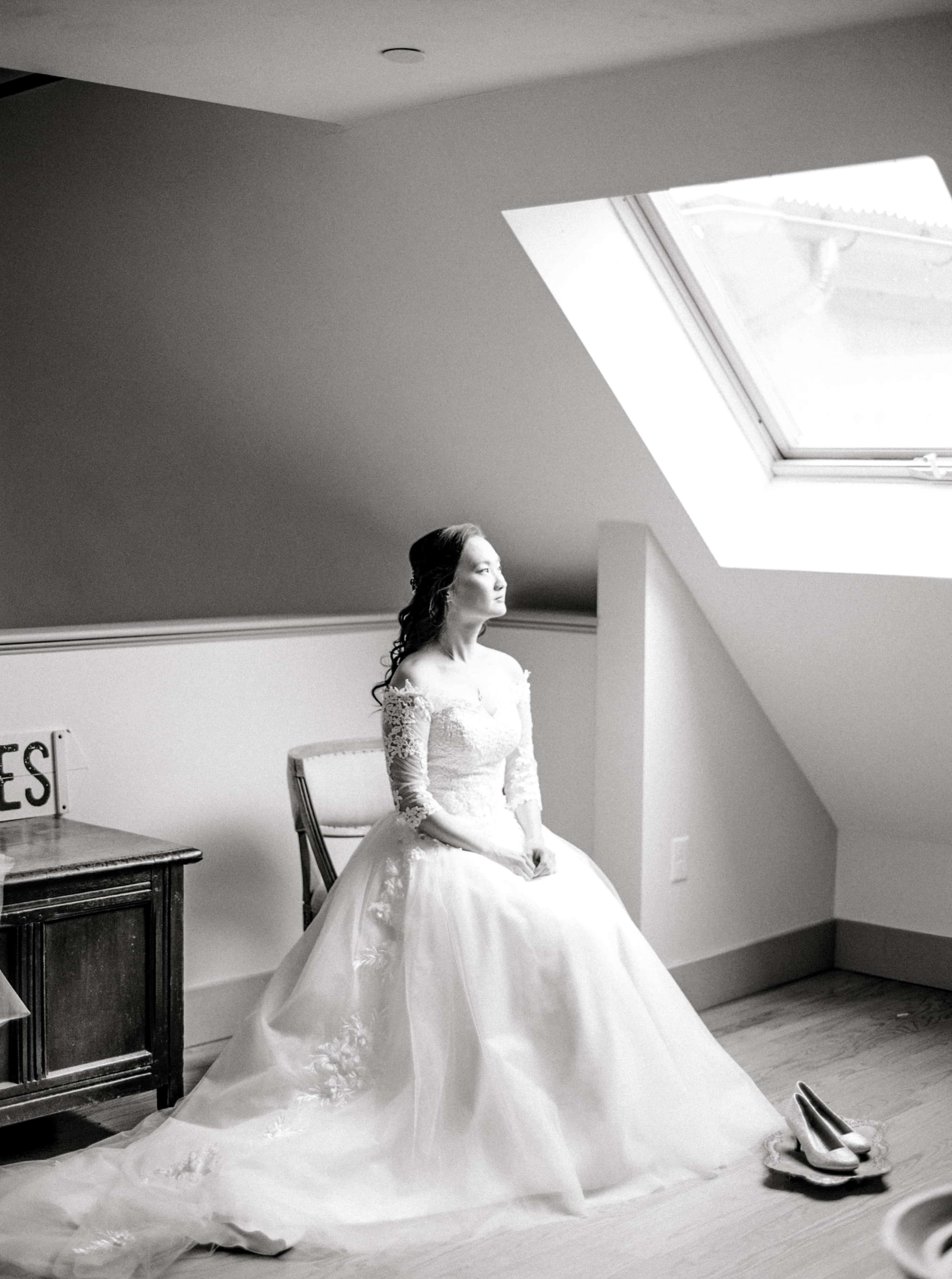 A bride is sitting in front of a large window feeling peaceful and stress-free before her wedding ceremony begins, photographed by North Shore, Massachusetts wedding photographer Marcela Plosker