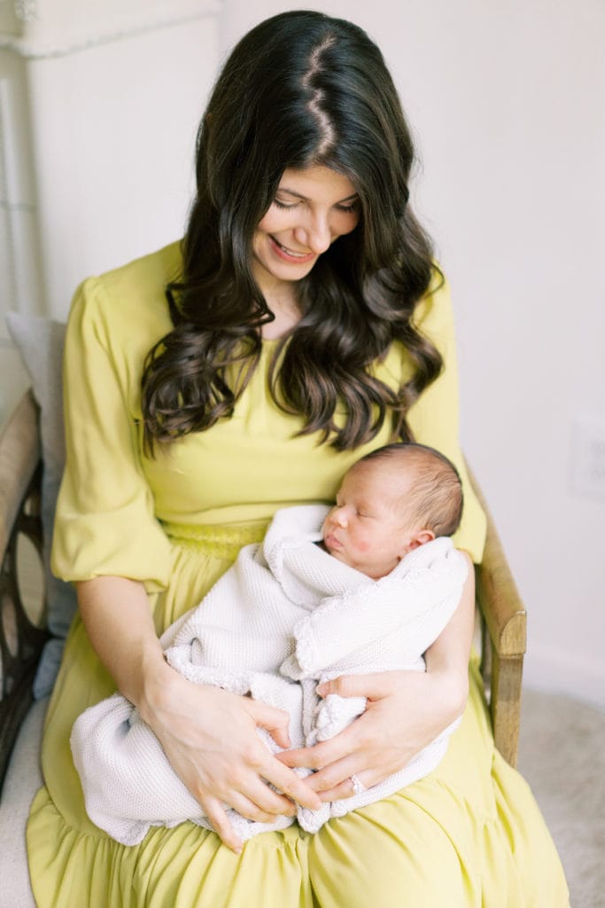 a woman in a yellow dress sit in a chair smiling down at her new born baby swaddled in a blanket