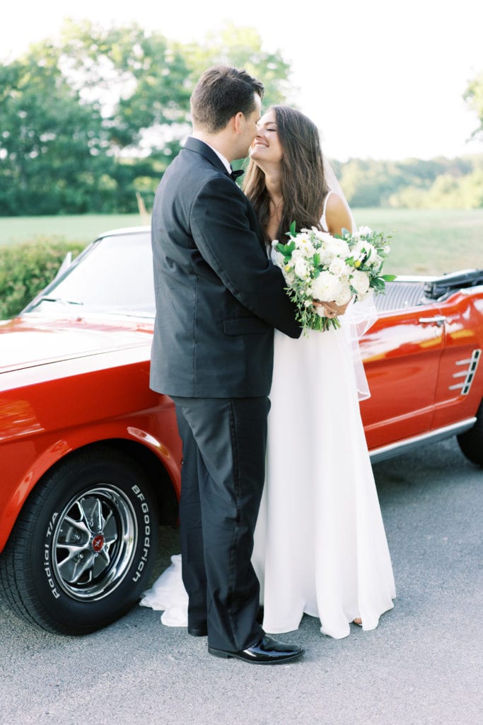 A bride and groom stand in front of a red vintage getaway car,  Photographed by North Shore, Massachusetts wedding photographer Marcela Plosker