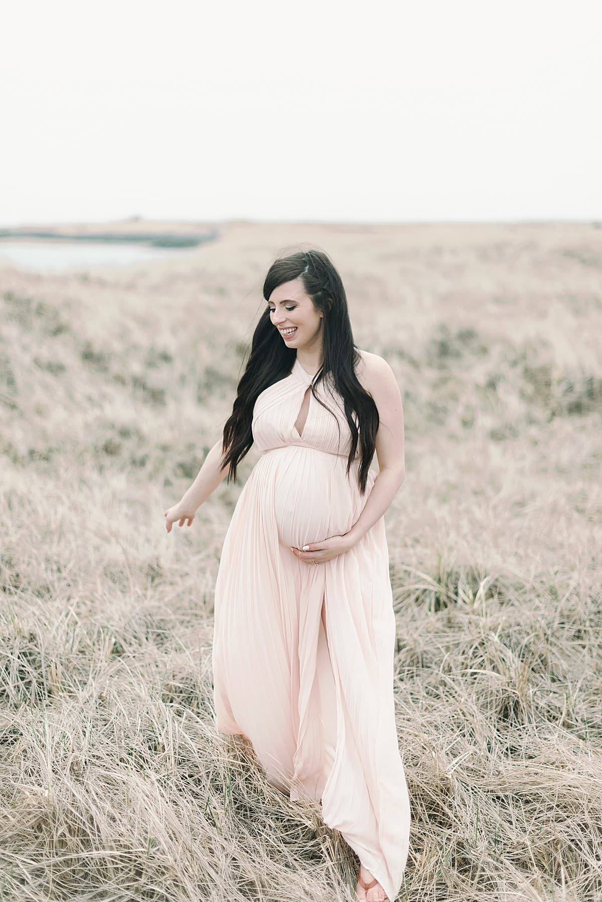 A pregnant woman stands in a grassy field in a blush dress holding her baby bump as she looks to the side smiling for a Mompreneur Guide to Creating a Mindful Morning Routine