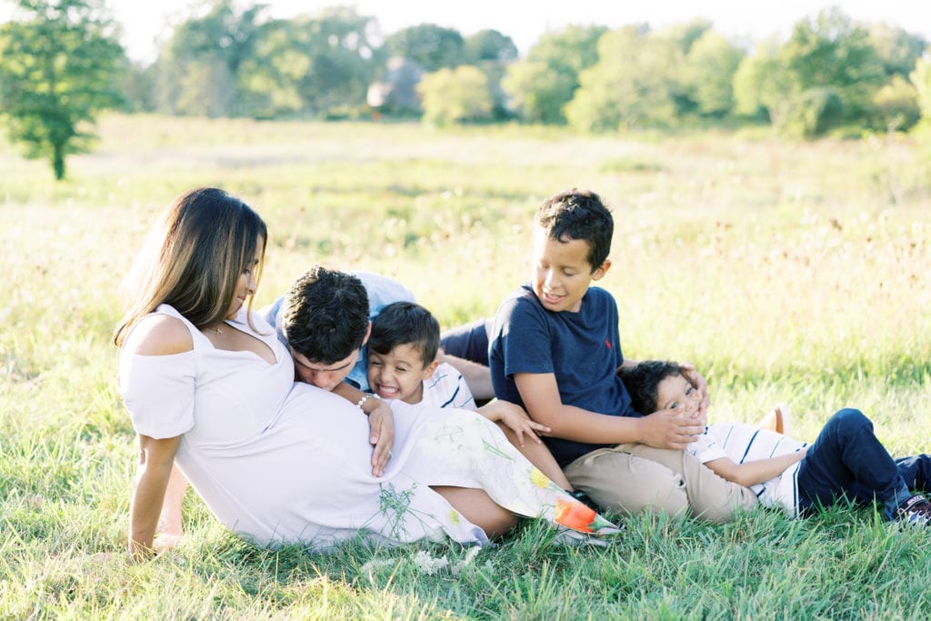 A mom laying in the grass surrounded by her children as her husband kisses her belly. Photographed by North Shore, Massachusetts wedding photographer Marcela Plosker