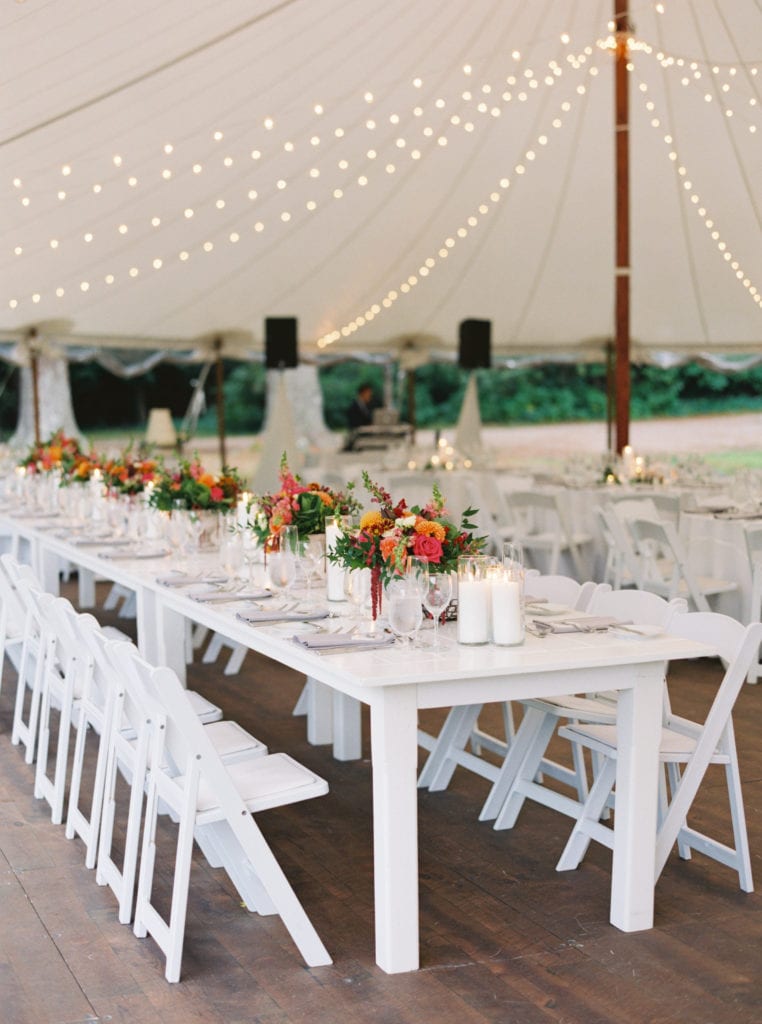A long white table with white chairs is set with wedding decor and wedding flowers underneath a white tent with strung lights. Photographed by Boston wedding photographer Marcela Plosker