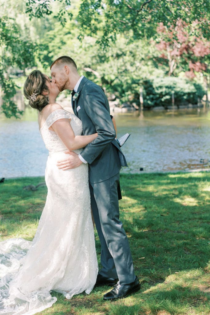 Newlywed couple embraces and kisses on the banks of a river in Boston, Massachusetts to celebrate their elopement. Photograph by Northshore, Massachusetts and Boston wedding photographer Marcela Plosker.