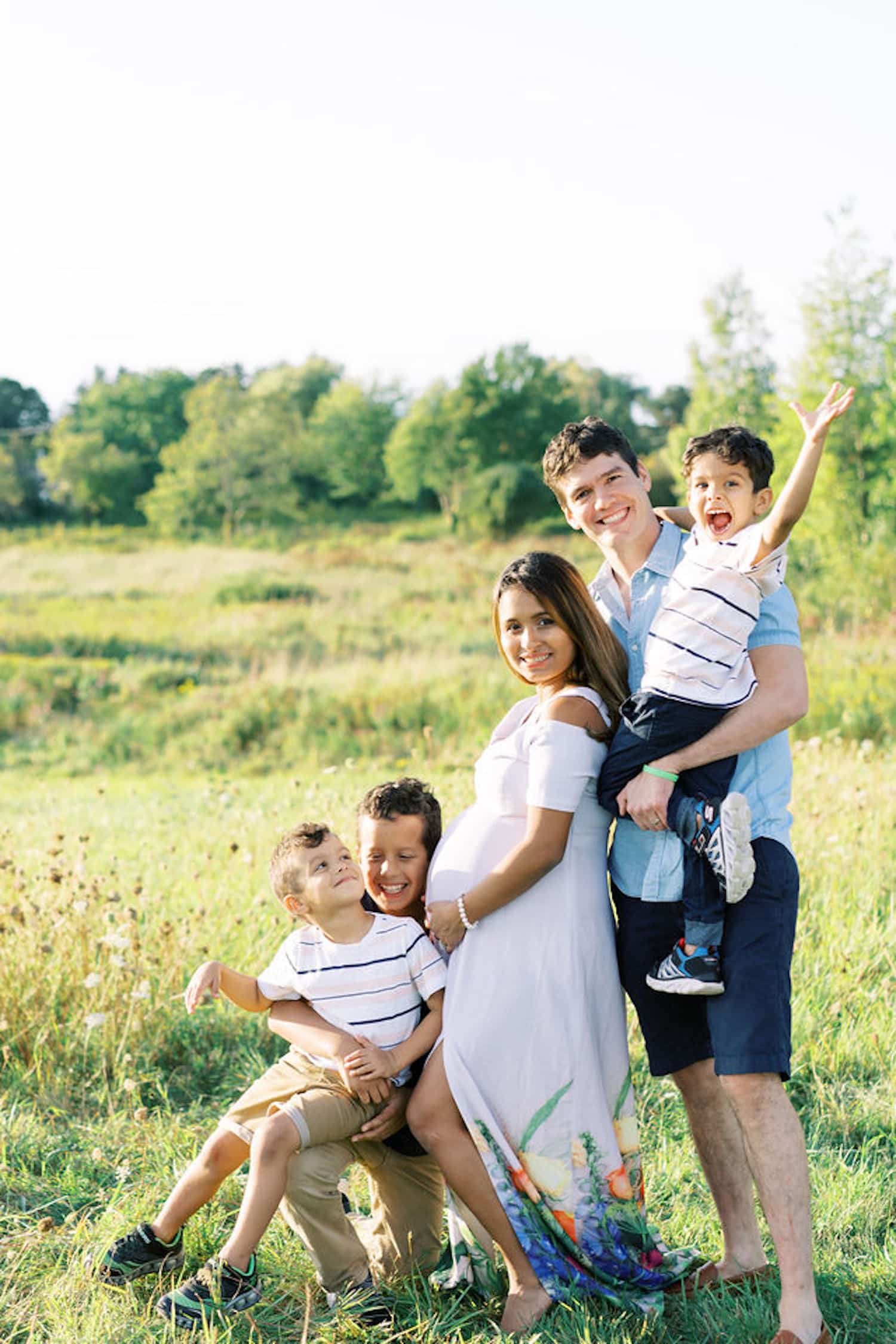 A pregnant mom smiles with her husband and three sons as they pose for a family photo in a field to celebrate The Benefits of Outsourcing as a Mompreneur.