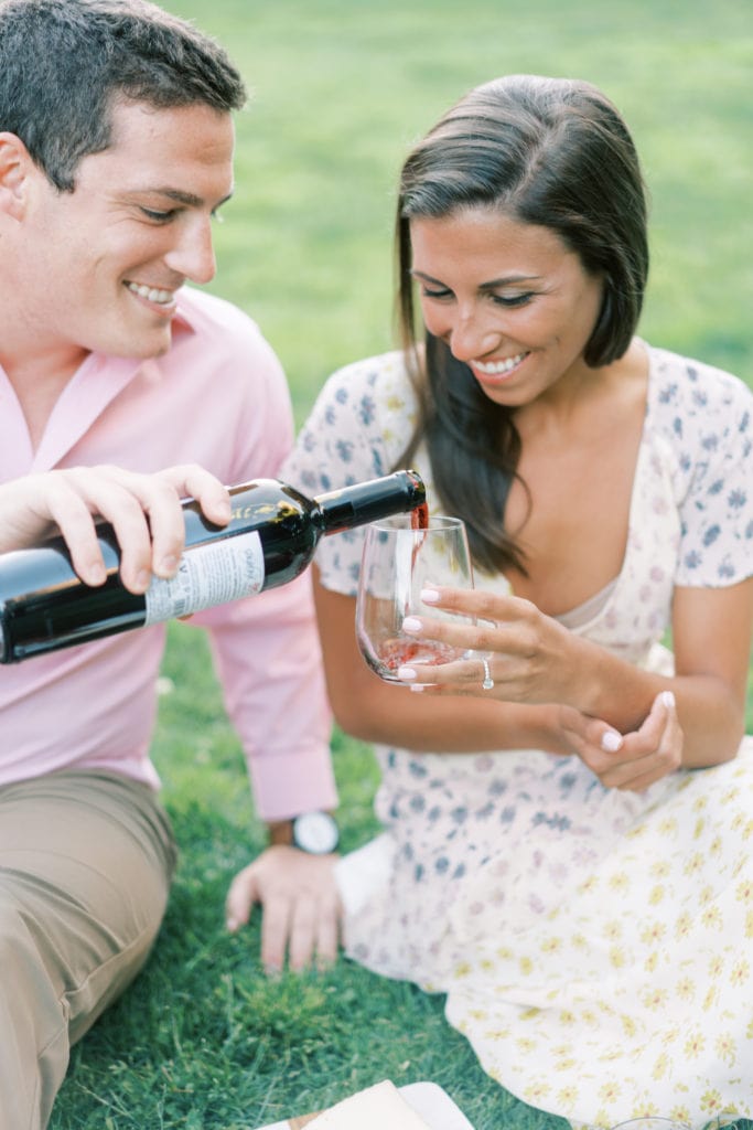 A groom to be pours his bride to be a glass of red wine during their engagement shoot. Image by Boston wedding photographer Marcela Plosker as an example of fine art engagement photo ideas.