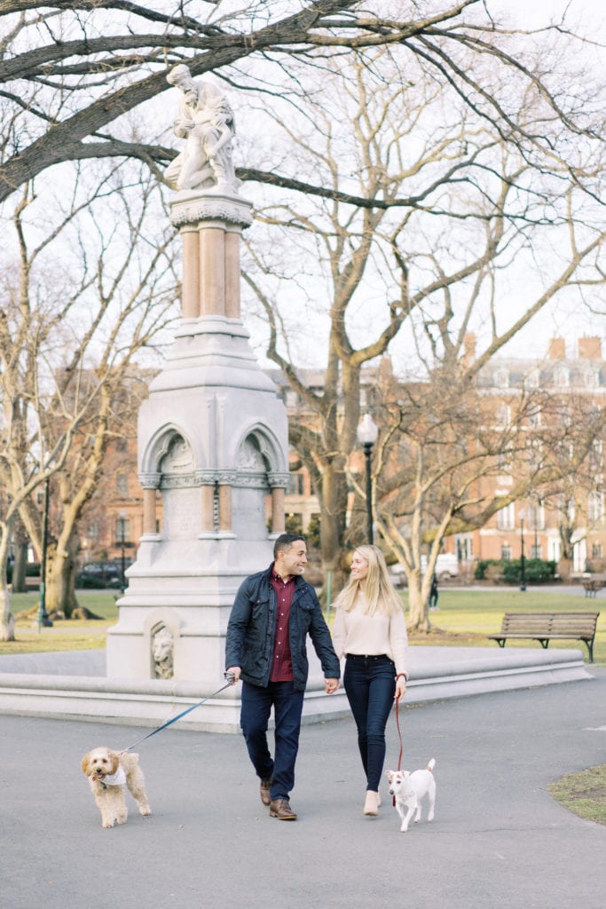A bride to be and groom to be hold hands while smiling at each other and walking their dogs in the park as an example of engagement photo ideas. Image by Marcela Plosker, a Massachusetts wedding and engagement photographer.