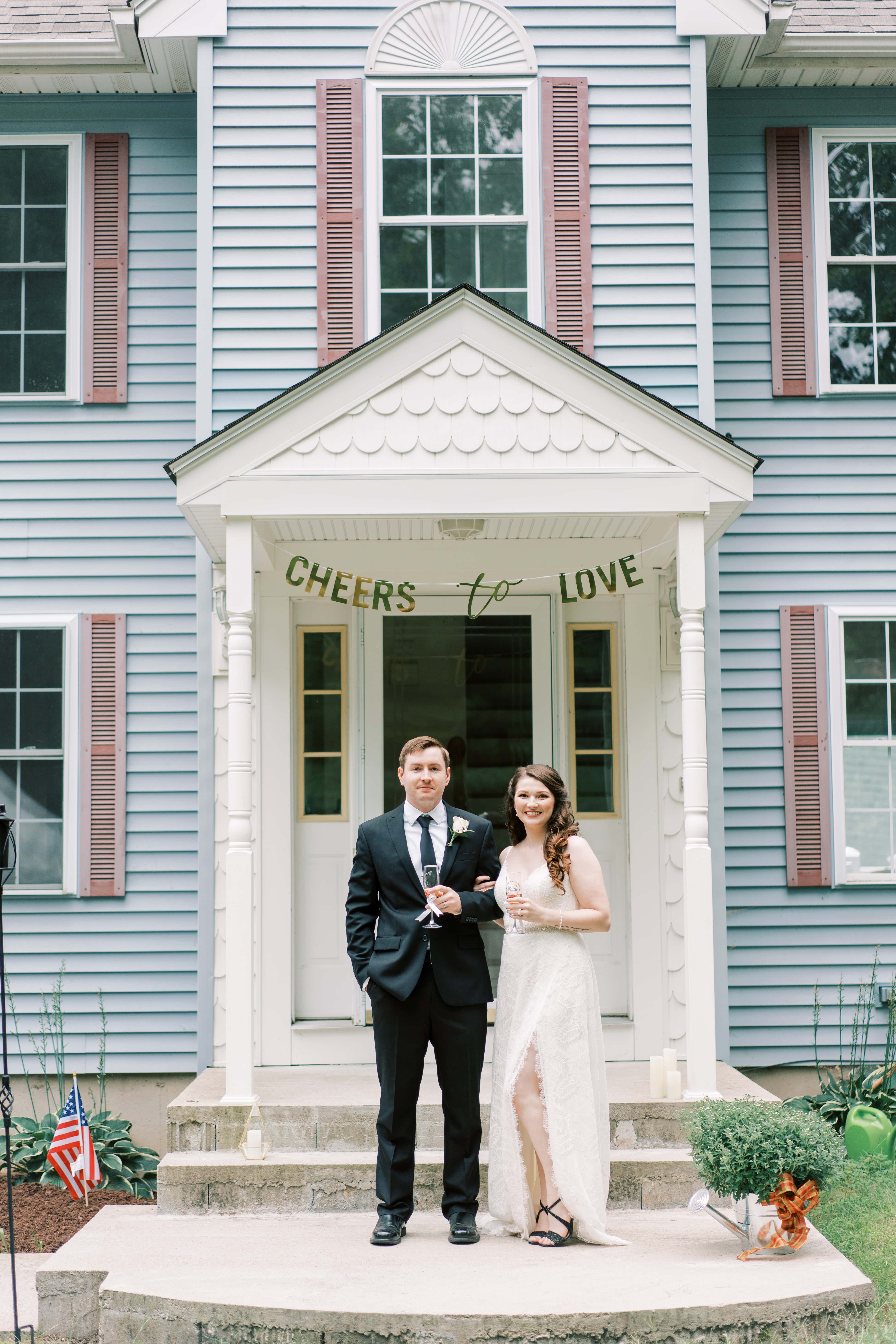 Bride and groom posing in front of their intimate small wedding venue with a banner hanging behind them that reads Cheers to Love
