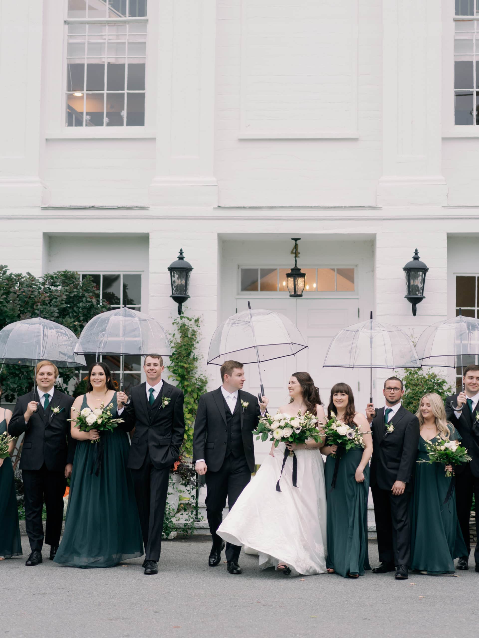Bride and groom pose with their entourage beside them, with all of them standing in pairs and holding a clear umbrella, shot by Marcela Diaz Photography